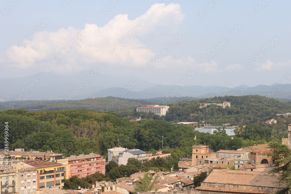 Fototapeta premium Panoramic view of Girona, Catalonia, Spain, featuring historic rooftops, lush green landscapes and distant mountain ranges under dramatic clouds. Scenic urban and natural landscape. 