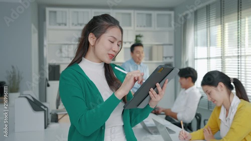 Female business professional using a digital tablet and stylus while working with colleagues in a modern office. The scene represents digital technology, workplace collaboration, productivity and prof