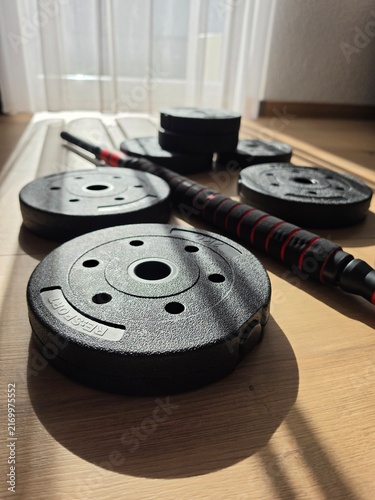 Obraz na płótnie Adjustable male dumbbell set with black weight plates and barbell connector bar on wooden floor in home gym illuminated by warm sunlight with soft curtain background and sharp linear shadows