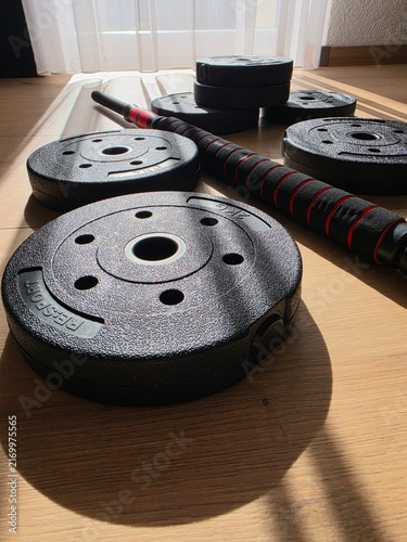 Quadro su tela Adjustable male dumbbell set with black weight plates and barbell connector bar on wooden floor in home gym illuminated by warm sunlight with soft curtain background and sharp linear shadows