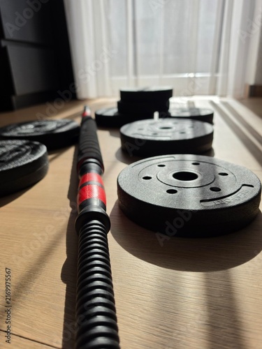 Konstfotografi Adjustable male dumbbell set with black weight plates and barbell connector bar on wooden floor in home gym illuminated by warm sunlight with soft curtain background and sharp linear shadows
