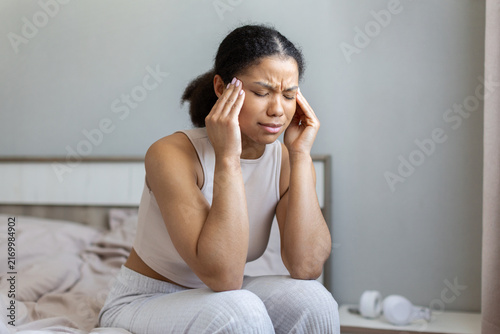 Foto Young Black woman sits on a bed with eyes closed and presses her temples, experi