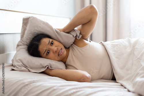 Fotografie Frustrated young Black woman lying in bed covering her ears with pillow, appeari
