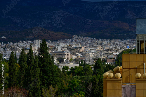 Elevated view overlooking the dense urban landscape of Athens, Greece, framed by Mediterranean trees and architecture with dramatic mountains rising in the background.