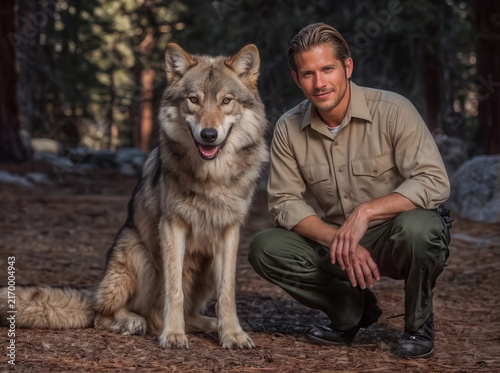 National park ranger sitting next to the gray wolf on forest background. Veterinary medicine, environmental protection and wildlife conservation concept. Lifestyle portrait for poster, visual material