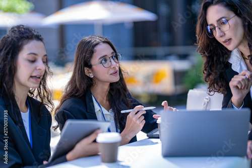 Bild auf Leinwand A diverse team of three Hispanic businesswomen in dark suits, deep in an animated discussion, sketch out an idea with a stylus on a tablet, part of a professional teamwork series