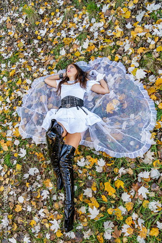 Woman wearing a gothic white dress and black leather boots lying among colorful autumn leaves and grass in an outdoor forest setting