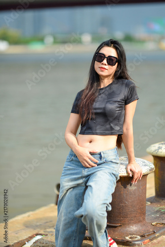Fashionable young Asian woman in sunglasses, black crop top and blue jeans leaning on rusty mooring bollard at riverside pier