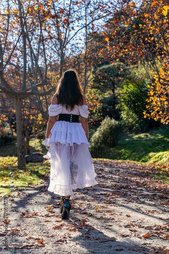 Woman wearing a white gothic dress, black corset and boots walking along a forest path surrounded by autumn foliage and golden leaves