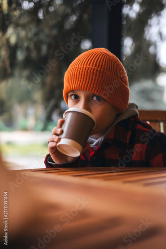 Young boy in a warm orange beanie drinking a hot beverage from a paper cup while sitting at a wooden table in a cozy cafe. Relaxing autumn lifestyle moment with a warm atmosphere