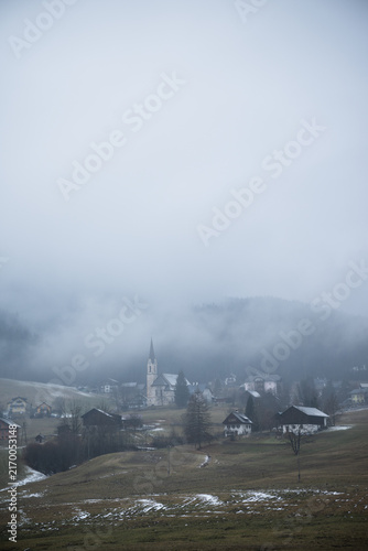 Vertical shot of foggy alpine village with tall church spire surrounded by mist covered hills and light snow in winter. Atmospheric winter landscape in Gosau, Austria.