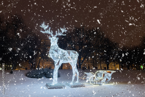 Glowing LED reindeer sculpture and sleigh in park during falling snow. Magical Christmas light decoration on snowy winter night, outdoor Christmas light.