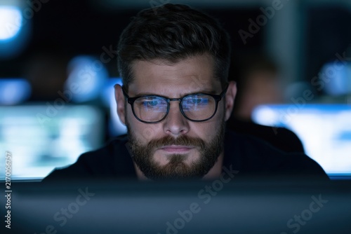Bearded man wearing glasses working late on computer screens