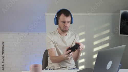 man playing video game in modern indoor room with laptop and controller, engaged and focused on screen, white walls and black furniture, plastic laptop and textile shirt.