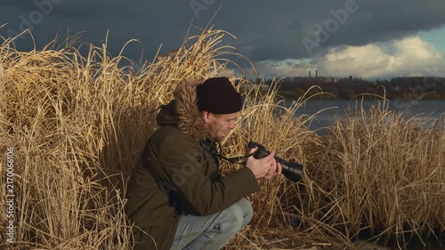 man holding camera in tall grass field, focused and concentrated on wildlife observation in nature photography setting with water body in background under cloudy sky.
