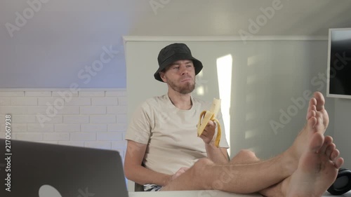 man in a hat posing with legs spread apart and hands on feet in an indoor setting, possibly an office or studio, neutral mood, white shirt and black hat, textile shirt, fabric hat, plastic.