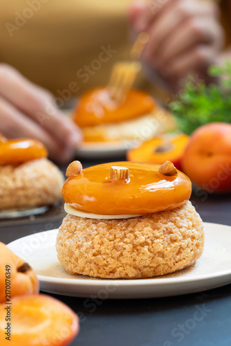 French apricot peach choux au craquelin pastry with owith shiny apricot and peach glaze and almond topping served on a white plate with person eating in background.