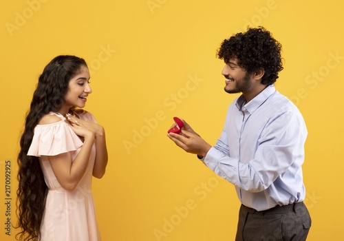 Bild auf Leinwand Surprised And Happy Young Hispanic Female And Male Couple Engaging In Romantic Proposal Moment, Holding Engagement Ring In Yellow Studio, Celebrating Love And Commitment With Joy