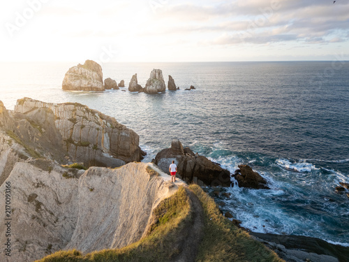 Aerial view of a man standing on a cliff edge overlooking the rugged coastline and dramatic rock formations in the sea at sunset in Liencres, Cantabria, Spain.