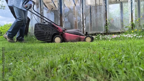 Spring lawn mowing. A young man in jeans mows the grass with a lawnmower in his backyard. Side view. 