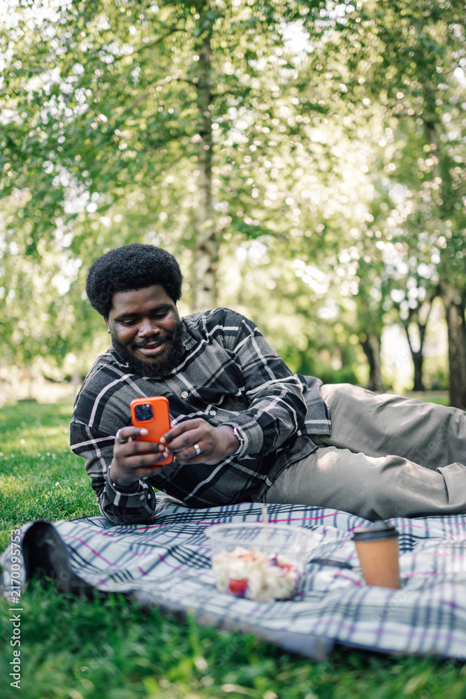 Fototapeta premium Young black man relaxing on picnic blanket in park, using smartphone, enjoying outdoor leisure and technology connection