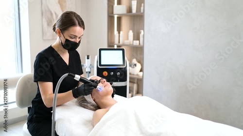 A professional cosmetologist wearing a medical mask, face shield, and gloves performs lip augmentation injections with a syringe for a young female client in a modern aesthetic medicine clinic