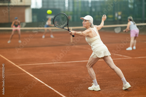 Fotografía Rear view of emotional aged woman enthusiastically playing tennis outdoors in summer, preparing to hit forehand to return ball to opponent court during doubles match with female partner