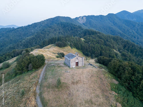 Aerial view of Parco Nazionale dell'Appennino Tosco-Emiliano featuring a historic stone chapel on a grassy ridge surrounded by dense forests, Provincia di Massa e, Italy.