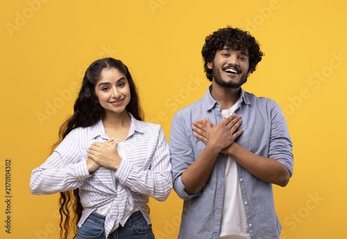 Fotografie Happy Young South Asian Couple Smiling With Hands On Their Chests, Standing Together With Joyful Expressions Against A Bright Yellow Background
