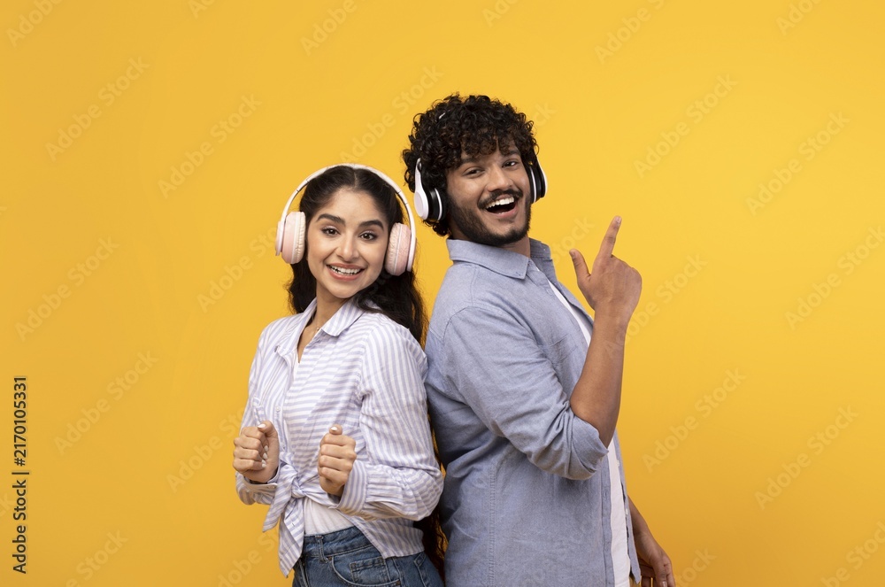 Fototapeta premium Smiling Young Asian Woman And Man Enjoying Music While Wearing Headphones, Standing Back To Back In Studio With Bright Yellow Background. Cheerful Emotion Captured In Fun Atmosphere.