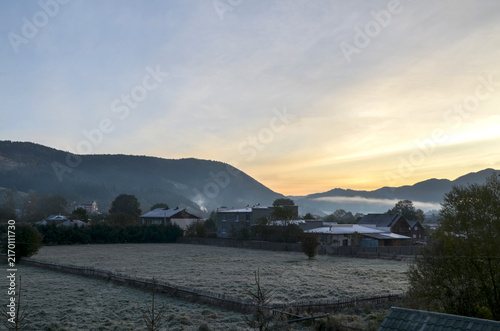 Soft morning mist drifts through a small rural village and frost-covered fields as the sun rises behind rolling mountains, creating a serene, peaceful dawn atmosphere over the countryside. Carpathians