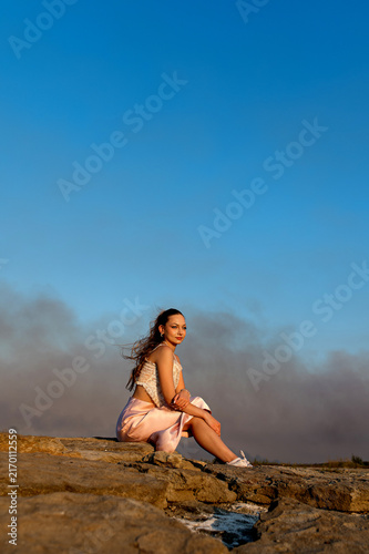 Photography Young woman sitting on rocky shore of the Dnipro River in Zaporizhzhia, Ukraine,  while heavy smoke from a large fire rises across the river