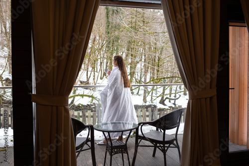 A young woman with long chestnut hair standing on a balcony of a country house, wrapped in a large white blanket, drinking tea from a mug, with a snowy winter forest in the background.
