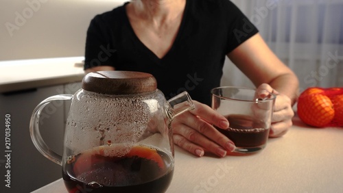 A woman sits at a table, holding a glass of coffee as steam rises from a nearby coffee pot. Oranges are placed on the table, adding color to the cozy scene.