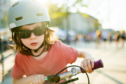 Cool young boy wearing helmet and sunglasses rides a kick scooter in the city on a sunny day. Active urban childhood, outdoor fun and safe mobility.