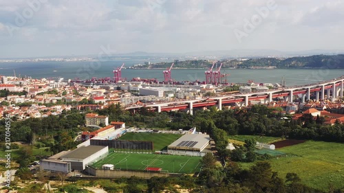 Drone flies over the industrial harbor and waterfront district of Lisbon, Portugal, with cargo terminals and the 25 de Abril Bridge visible in the distance.