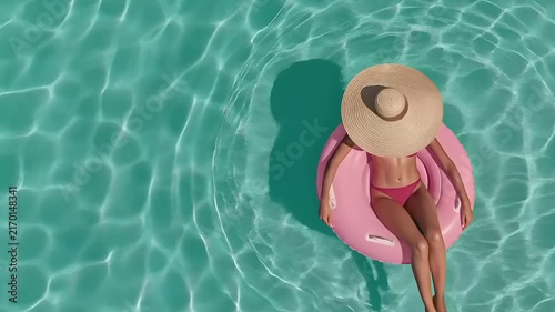 Summer hot vibe lifestyle. A topdown view of a person floating on a pink inflatable ring in a turquoise swimming pool. The person wears a widebrimmed straw hat and a pink bikini.