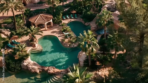 Summer hot vibe lifestyle. An aerial view of a luxurious resort features a large, turquoise swimming pool surrounded by lush greenery and towering palm trees.