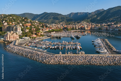 Aerial view of the harbor filled with numerous yachts and boats, protected by a stone breakwater, with the coastal town and green hills in the background in Rapallo, Liguria, Italy.