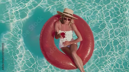 Summer hot vibe lifestyle. A vibrant, topdown view of a woman floating on a red inflatable ring in a swimming pool. She wears a white swimsuit, a widebrimmed hat, and sunglasses.
