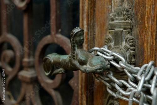 Close-up of vintage ornate bronze door handle shaped like female figure wrapped with metal chain on old wooden door