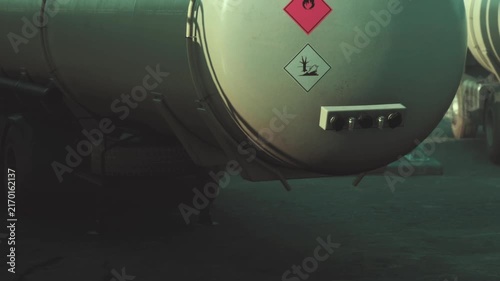 A tanker truck is parked in an industrial location. The truck has warning labels indicating hazardous materials. Nearby vehicles and equipment are visible in the background.
