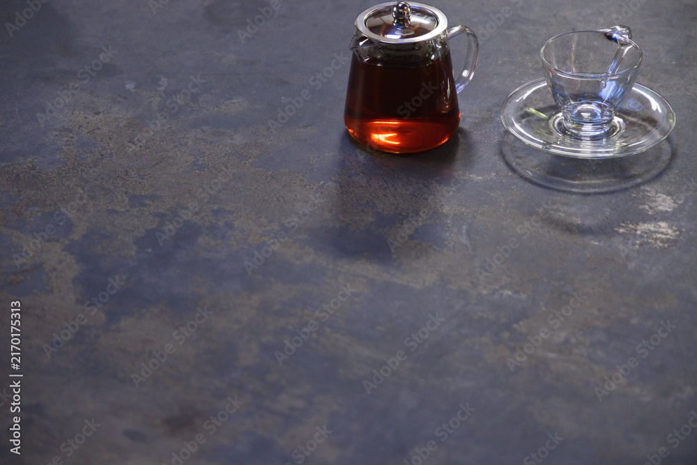 Fototapeta premium Clear glass teapot with metal lid and teacup on saucer resting on textured tabletop, copy space