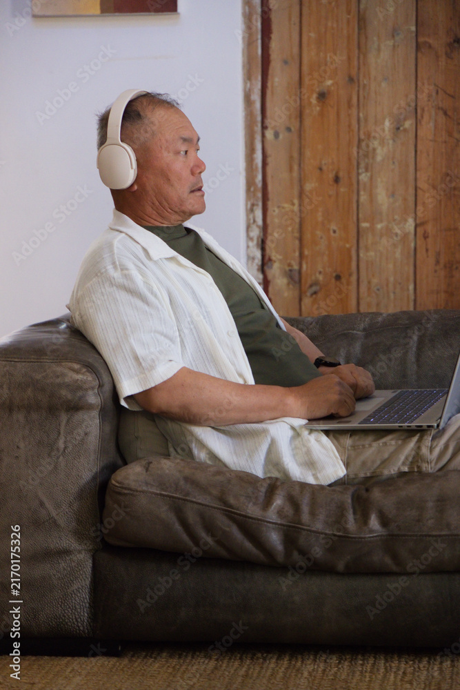 Fototapeta premium Senior Asian man sitting on dark brown leather couch wearing headphones and typing on laptop