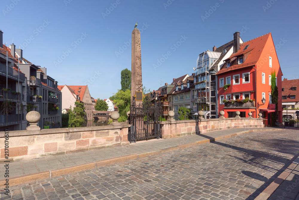 Fototapeta premium Stone monument beside the Pegnitz River surrounded by traditional buildings and cobblestone streets in Nuremberg, Germany.