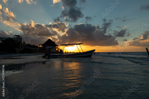 Картината върху платно Small fishing boat silhouette on calm beach shallow water under sun breaking through dark clouds