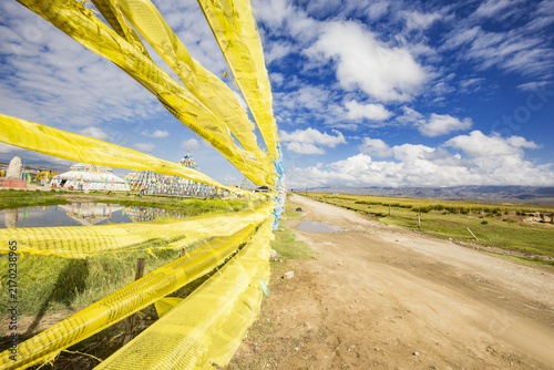 Quadro su tela Bright yellow prayer flags stretch along a dirt road in an open grassland, under a blue sky with white clouds