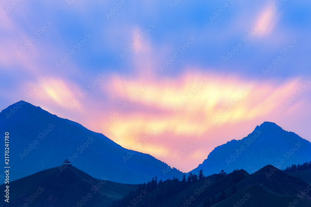 Obraz premium Dark mountain silhouettes stand against a vibrant twilight sky with soft colored clouds, creating a serene natural landscape.