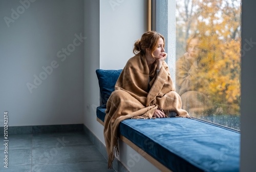 Pensive young woman wrapped in a warm blanket sits on a window sill looking outside on a rainy autumn day