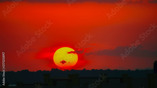 Bild auf Leinwand A striking sunset view featuring a vivid glowing red and orange sun dipping below dark cloud layers over a distant horizon